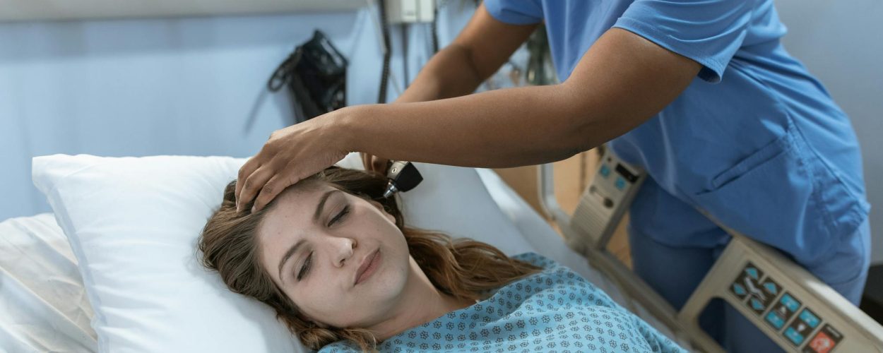 Crop African American female doctor with professional equipment doing examination of ear of woman lying on bed in hospital ward